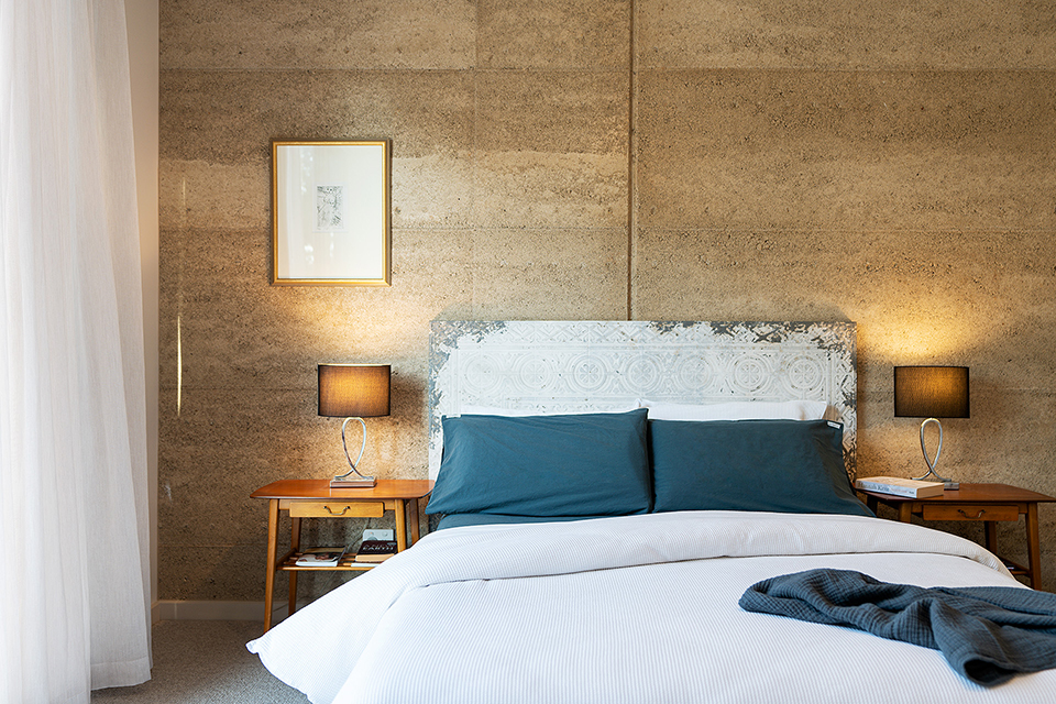 Antique bedhead and mid-century bedside tables with rammed earth wall behind bed