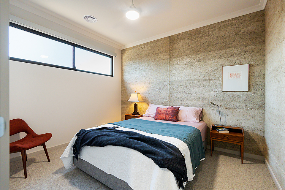 Side on view of bedroom with mid-century bedside tables and a rammed earth wall behind the bed